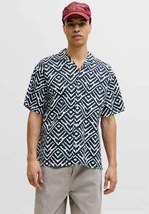 Homme portant une chemise à manches courtes noire et blanche à motif géométrique, un pantalon beige et une casquette rouge avec un texte.