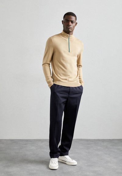 Beige zippered trøje med høj krave, parret med marineblå bukser og hvide sneakers, der har en glat tekstur og tætsiddende design.