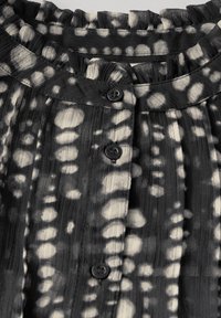 Schwarz-weiße Bluse mit Rüsche am Ausschnitt, mit Knopfverschluss und strukturierter Stoff mit einem kreisförmigen Muster.