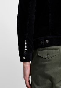 Personne portant une veste en velours côtelé noire avec boutons-pression et un pantalon vert olive avec une poche arrière boutonnée, main détendue sur le côté.