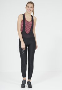 Zwarte fietsbibs met reflecterende accenten en een aansluitend ontwerp, gecombineerd met een bordeauxrood aansluitend tanktop. Het materiaal lijkt rekbaar en ademend.