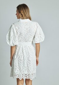 Biała sukienka z koronki o kwiatowym wzorze, z bufiastymi rękawami, kołnierzykowym dekoltem i zebranym w talii, sięgająca powyżej kolana.