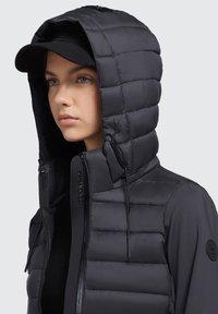 Čierna prešívaná bunda s veľkou kapucňou, ktorá má prešívanú štruktúru, predný zips a nastaviteľné šnúrky. Akcentové logo na rukáve.