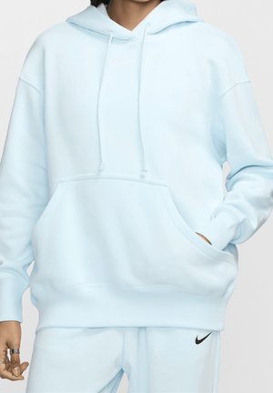 Person wearing light blue Nike hoodie and matching sweatpants, with hand partly in hoodie pocket and rings on fingers.