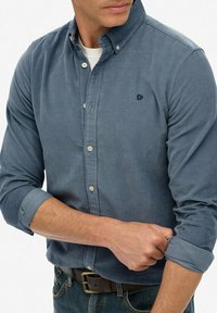 Uomo che indossa una camicia azzurra con bottoni, arrotola le maniche, con una maglietta bianca visibile, jeans blu e una cintura marrone, su uno sfondo semplice.