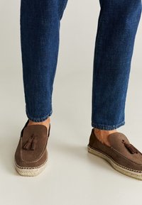 Gambe che indossano jeans blu scuro e mocassini con nappine marroni e suola in espadrilles intrecciata, in piedi su un pavimento chiaro e neutro.