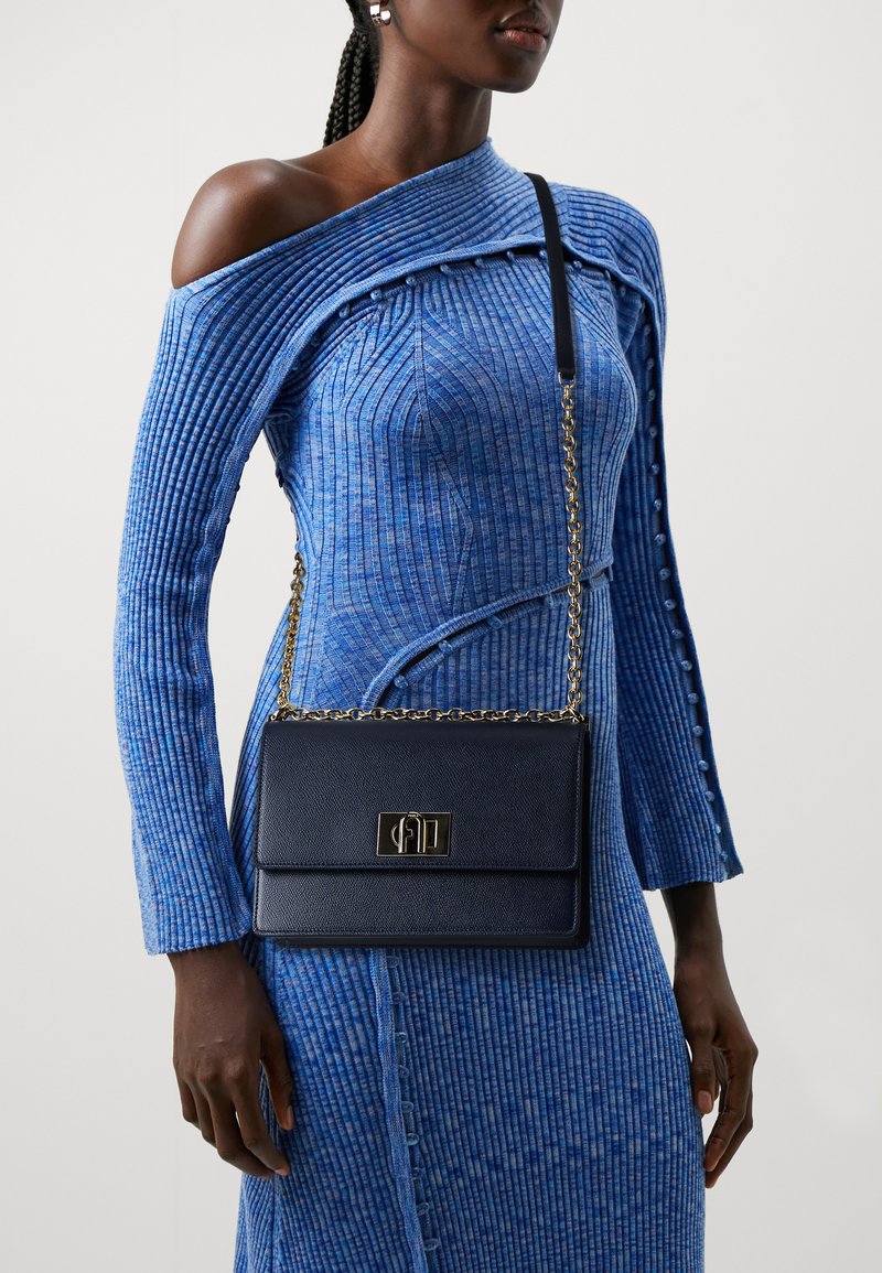 Borsa a tracolla in pelle blu navy con chiusura in hardware dorato, caratterizzata da un design strutturato e una tracolla a catena, indossata con un vestito blu a coste.