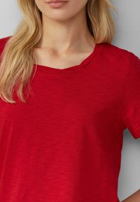 Blonde vrouw draagt een heathered rode t-shirt met korte mouwen en een ronde halslijn, afgekort bij de hals en het bovenlichaam.