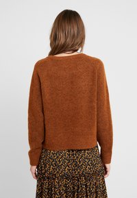 Brązowy, puszysty sweter z dzianiny z okrągłym dekoltem i długimi rękawami, prezentowany na tle czarniej spódnicy w kwiatowy wzór. Miękka tekstura, luźny krój.