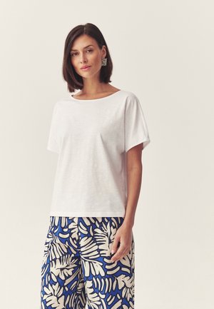 Woman with shoulder-length brown hair wearing square silver earrings, a plain white short-sleeve top, and blue-and-white patterned wide-leg pants.