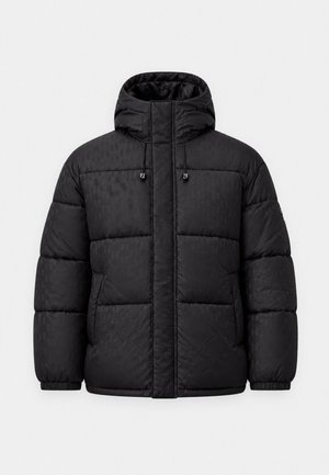 Jeune homme portant une veste matelassée noire avec capuche, un polo noir, un pantalon noir et des baskets noires à semelles blanches, debout devant un fond blanc.
