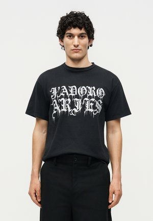 Jeune homme aux cheveux courts, bouclés et foncés, portant un t-shirt noir avec un texte blanc de style gothique « JADORE ARIES » sur un fond clair uni.