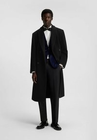 Cappotto in lana nera su un gilet in velluto blu navy, camicia bianca e pantaloni eleganti neri. Scarpe eleganti nere e lucide completano il look.
