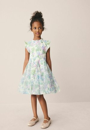 Junges Mädchen mit lockigem Haar, das ein pastellfarbenes Blumenmusterkleid und beige glitzernde Ballerinas trägt, vor einem neutralen Hintergrund steht.