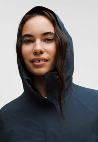 Jachetă impermeabilă navy, cu guler înalt și glugă ajustabilă, având un fermoar frontal și o textură mată, netedă.