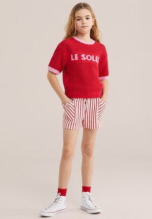 Mladi djevojka s dugom kosom nosi crvenu pletenu majicu s natpisom "LE SOLEIL", crvene i bijele prugaste kratke hlače, crvene čarape i bijele tenisice visokog gazišta.