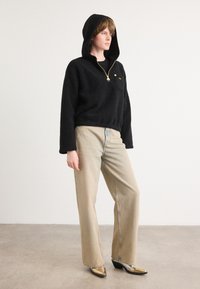 Pessoa vestindo um fleece preto com capuz, jeans beige de perna larga e botas douradas metálicas de bico fino, em pé sobre um chão cinza com fundo branco.