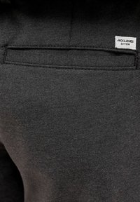 Nahaufnahme der hinteren Tasche einer dunklen Stoffhose mit einem kleinen weißen Etikett, auf dem "JACK & JONES EST. 1990" oberhalb der Tasche aufgenäht ist.