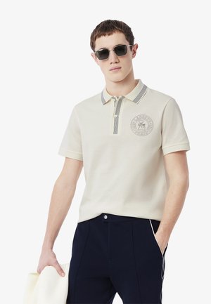Beige pikétröja med randig krage, med en logotyp på bröstet och korta ärmar. Matchad med mörkblå byxor.