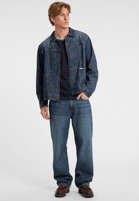 Giubbotto in denim blu scuro con chiusura a bottoni, due tasche sul petto e una vestibilità morbida. Abbinato a una camicia blu scuro e jeans in denim a gamba larga.