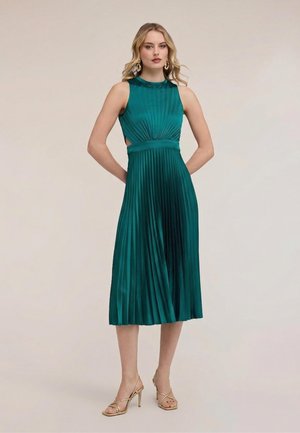 Mujer de pie con las manos detrás de la espalda, vistiendo un vestido midi plisado sin mangas verde esmeralda y sandalias doradas de tacón alto.