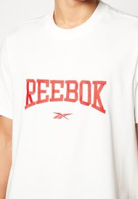 Vit t-shirt med en röd broderad "REEBOK"-logotyp och en mindre logotyp nedanför. Har en klassisk rund halsringning och korta ärmar.