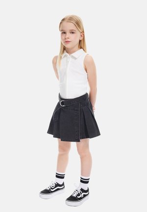 Junges Mädchen mit langen blonden Haaren, das ein ärmelloses weißes Zip-Up-Top, einen schwarzen plissierten Rock mit Gürtel, gestreifte Socken und schwarze Turnschuhe trägt und steht.
