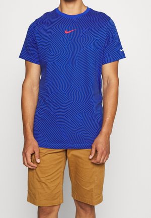 Homme portant un t-shirt Nike bleu à motifs avec un logo rouge et un short beige, debout les bras détendus le long du corps sur fond blanc.