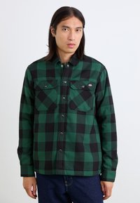 Chemise à carreaux verte et noire, en tissu brossé, avec deux poches poitrine, des boutons pression et un col classique.