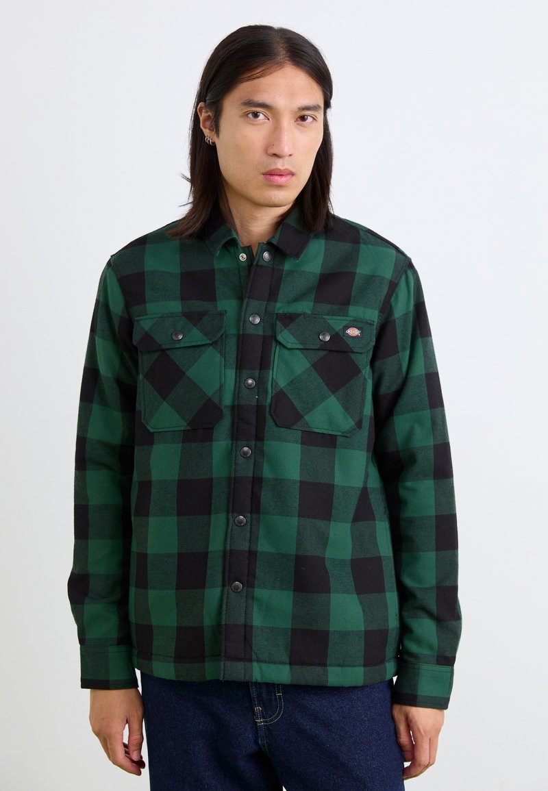 Chemise à carreaux verte et noire, en tissu brossé, avec deux poches poitrine, des boutons pression et un col classique.
