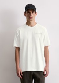 White cotton t-shirt with a relaxed fit and short sleeves, featuring subtle text near the chest. Paired with dark trousers and a black cap.