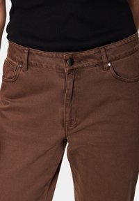 Rjavi denim jeans z gladko teksturo, ki imajo klasičen pet-žepni dizajn, zapiranje na gumb in srebrne kovinske dodatke.