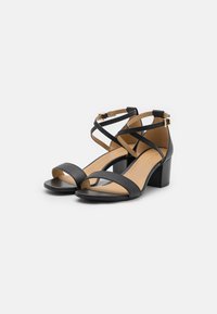 Sandales noires à talons avec des brides croisées et un talon carré. Fabriquées en cuir lisse avec une semelle intérieure beige. Design à bout ouvert, style minimaliste.