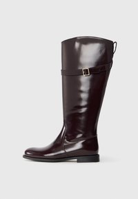 Bota marrón brillante hasta la rodilla con punta redondeada, suela plana y una correa decorativa con un hebilla de metal cerca del tobillo. Textura suave.