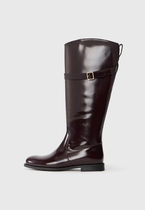 Botte marron brillante jusqu'au genou avec un bout arrondi, une semelle plate et une sangle décorative dotée d'une boucle en métal près de la cheville. Texture lisse.