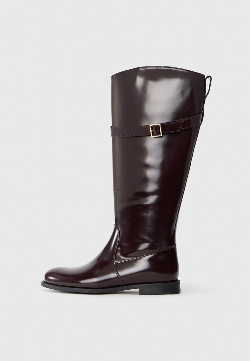 Bota marrón brillante hasta la rodilla con punta redondeada, suela plana y una correa decorativa con un hebilla de metal cerca del tobillo. Textura suave.
