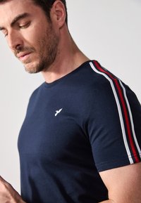 Camiseta de algodón azul marino con franjas blancas y rojas en los hombros, con un pequeño logo blanco en el pecho; cuello redondo, mangas cortas.