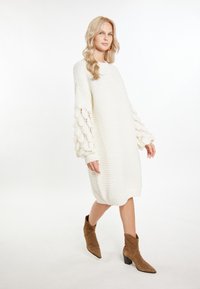 Abito a maglia crema con texture a coste e maniche a palloncino, caratterizzato da un motivo ondulato. Abbinato a stivaletti alla caviglia in suede marrone.