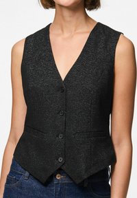 Kvinna som bär en ärmlös svart knäppt väst med subtil glans över blå denimjeans, avkortad nedanför axlarna.
