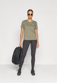 Camiseta verde oliva de manga corta con un pequeño logo, combinada con leggings negros, sosteniendo una mochila negra y usando gafas de sol.