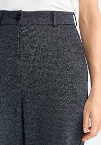Pantalones de tela texturizada gris oscuro con cierre de botón y trabillas para cinturón. Presentan un ajuste afinado y un ligero brillo en el material.