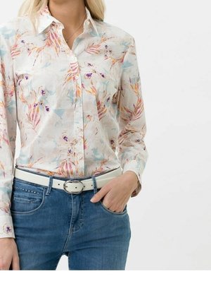 Vrouw draagt een pastelkleurig bloemenoverhemd, ingestopt in een blauwe spijkerbroek met een witte riem, één hand in de zak, tegen een effen achtergrond.