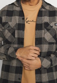 Svart och grå rutigt flanellskjorta lagd över en beige t-shirt med en svart signaturtryck, som visar knappar och bröstfickor.