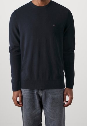 Mann trägt einen schlichten schwarzen Langarmpullover mit kleinem Logo auf der Brust und ausgewaschene graue Jeans, steht vor einem einfarbigen Hintergrund.