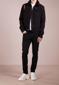 Giacca in denim nero con colletto in shearling, due tasche sul petto e chiusura frontale con bottoni. Indossata sopra una maglietta bianca e abbinata a pantaloni neri.