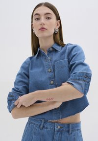 Džínová zkrácená košile s knoflíkovým zapínáním vpředu, klasickým límečkem a kapsou na prsou. Vlastnosti zahrnují ohrnuté rukávy a třepený lem.
