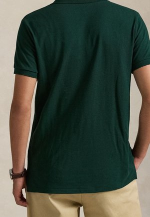 Mann trägt ein dunkelgrünes kurzärmeliges Polohemd und beige Hose, von hinten gezeigt, mit einer Armbanduhr am linken Handgelenk.