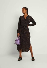 Mulher de pé com a mão na anca, vestindo um vestido midi de manga longa preto com padrão, sandálias pretas de salto alto e segurando uma pequena bolsa roxa.