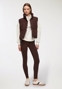 Gilet imbottito marrone con colletto alto, abbinato a una felpa bianca, leggings marroni e sneakers bianche con dettagli marroni.