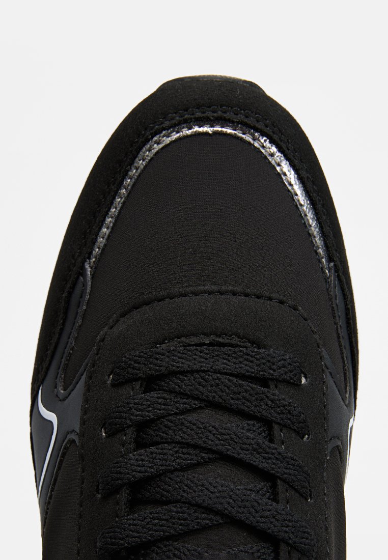 Zapatilla deportiva negra con parte superior de tela suave, cordones texturizados y un ribete metálico plateado a lo largo del borde. Diseño minimalista con puntera redondeada.